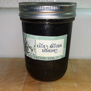 Könnte beinhalten: Ein Glasgefäß mit schwarzem Deckel und einem weißen Etikett mit der Aufschrift "Katie's Natural Remedies Elderberry Syrup".