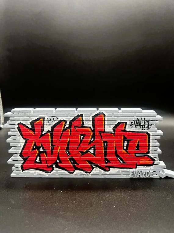 A miniature graffiti-tagged brick wall nameplate — 3D printed, hand painted in street-art style, it looks like someone tagged a tiny alley just for you.