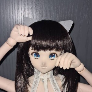 Könnte beinhalten: Eine Puppe mit langen schwarzen Haaren, die ein weißes Oberteil mit silbernen Akzenten und einen weißen Rock trägt. Die Puppe hat blaue Augen und trägt ein weißes Katzenohr-Haarband.