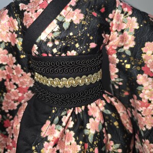 May include: A black kimono-style garment with a floral pattern in pink and gold. The garment features a black belt with gold trim and decorative black trim. The sleeves are long and the fabric has a slight sheen.