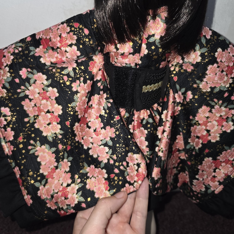 May include: A black kimono-style top with a floral pattern of pink cherry blossoms. The fabric has a subtle sheen, and the design includes a black waist cincher with gold accents. The garment has a ruffled hem.
