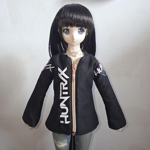 Könnte beinhalten: Eine schwarze Jacke mit dem Wort "HUNTRAX" in weißen Buchstaben auf der Vorderseite. Die Jacke hat einen roségoldenen Reißverschluss und weiße und blaue Akzente an den Ärmeln. Die Jacke wird von einer Puppe mit schwarzen Haaren und blauen Augen getragen.
