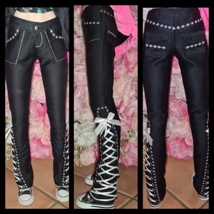 May include: Black denim trousers with white stitching and a lace-up design on the sides. The trousers have a button closure and pockets with decorative stitching. The bottom of the trousers are paired with black and white trainers.