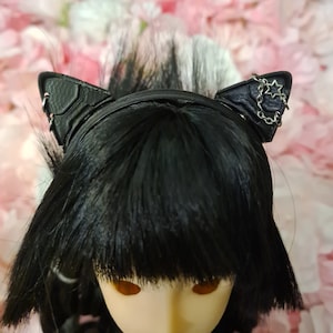 Könnte beinhalten: Eine schwarze Puppe mit schwarzen Haaren, die ein schwarzes Stirnband mit zwei schwarzen Katzenohren trägt. Die Katzenohren haben silberne Akzente und eine silberne Kette mit einem Sternanhänger.