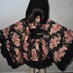 May include: A black floral kimono with pink blossoms and gold accents. The kimono features a black belt with gold trim and black ruffled edges. The garment is designed for a doll.