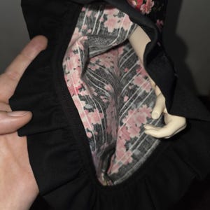 May include: Black dress with a ruffled hem, displaying a floral lining with pink blossoms on a black and white striped pattern. The dress is held open, revealing the interior design.