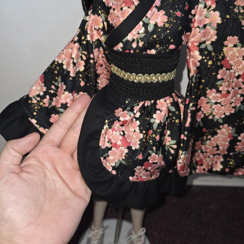 May include: A black kimono-style doll dress with a floral pattern in pink and gold. The dress has a black obi belt with gold trim and a ruffled hem. The sleeves are wide and flowing.
