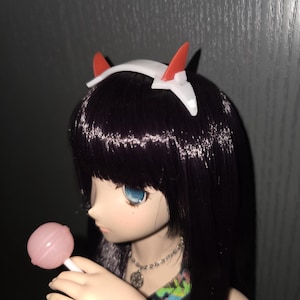 Könnte beinhalten: Eine Puppe mit schwarzen Haaren und blauen Augen, die ein weißes Stirnband mit roten und schwarzen Akzenten trägt. Die Puppe hält einen rosa Lutscher. Die Puppe trägt eine Halskette.