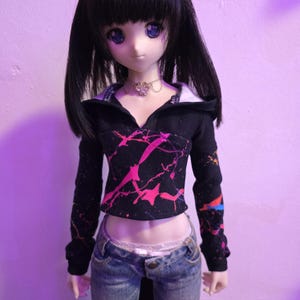 Könnte beinhalten: Eine Puppe trägt ein schwarzes, langärmeliges Crop-Top mit einem rosa und lila Spritzmuster. Die Puppe trägt außerdem eine blaue Jeans und eine silberne Halskette. Die Puppe hat schwarzes Haar und blaue Augen.