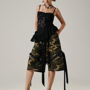 May include: A black lace top with spaghetti straps and a ruffled peplum is paired with camouflage print culottes. The culottes have black ribbon details and are worn with patterned heeled mules. A dark necklace completes the outfit.