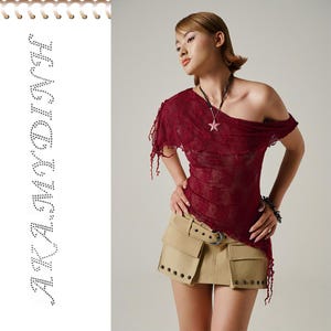 May include: A burgundy off-the-shoulder top with a star pendant necklace and a khaki mini skirt. The top has a sheer, lacy texture and fringed edges. The skirt features a belt with a silver buckle and multiple pockets. The word "AKAMDINH" is spelled out in rhinestones.