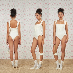 Peut inclure: Un body blanc en dentelle ajourée, avec des bords à volants et un décolleté carré. Le vêtement une pièce présente un détail de nœud à la poitrine. La modèle porte des chaussettes et des talons blancs, tenant une fleur blanche. Fond floral.