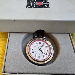 May include: A Swiss Army brand pocket watch with a white face, black Arabic numerals, and a red inner ring, housed in a grey presentation box. The watch features luminous hands and the Swiss Army logo on the dial and the box lid.