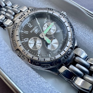 May include: A silver-tone Fossil Blue analog watch with a dark gray dial, featuring a stainless steel link bracelet, a rotating bezel with compass markings, luminous hands, and three sub-dials displaying the day, date, and a 24-hour cycle.