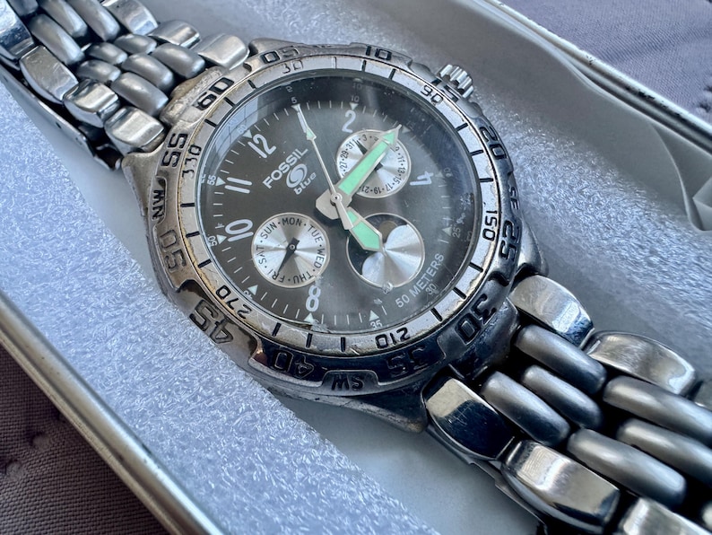 May include: A silver-tone Fossil Blue analog watch with a dark gray dial, featuring a stainless steel link bracelet, a rotating bezel with compass markings, luminous hands, and three sub-dials displaying the day, date, and a 24-hour cycle.