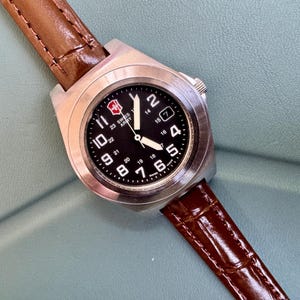 May include: A stainless steel Swiss Army watch with a black face and a brown leather band. The watch face displays white numerals and a red and white logo. The watch is angled across a light blue surface.