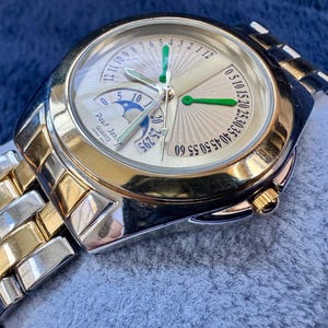 May include: A gold and silver-toned Paul Jar Quartz watch. The watch face is light gold with green and silver hands, and a moon phase indicator. The watch band is a combination of gold and silver links. The watch is displayed on a blue surface.