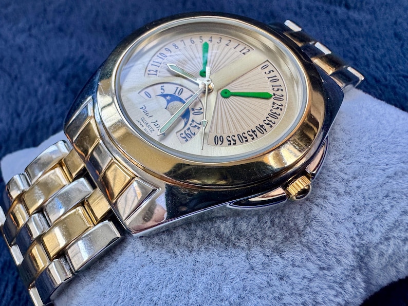 May include: A gold and silver-toned Paul Jar Quartz watch. The watch face is light gold with green and silver hands, and a moon phase indicator. The watch band is a combination of gold and silver links. The watch is displayed on a blue surface.