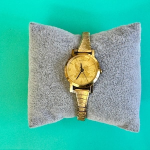 May include: A gold-toned wristwatch with a textured band and face, resting on a gray, plush pillow. The watch face features a detailed design. The watch is a classic accessory.