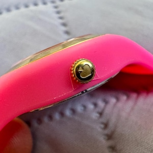 May include: Close-up of a bright pink wristwatch with a gold-colored bezel and crown. The crown features a heart-shaped design. The watch band is made of a flexible, pink material.