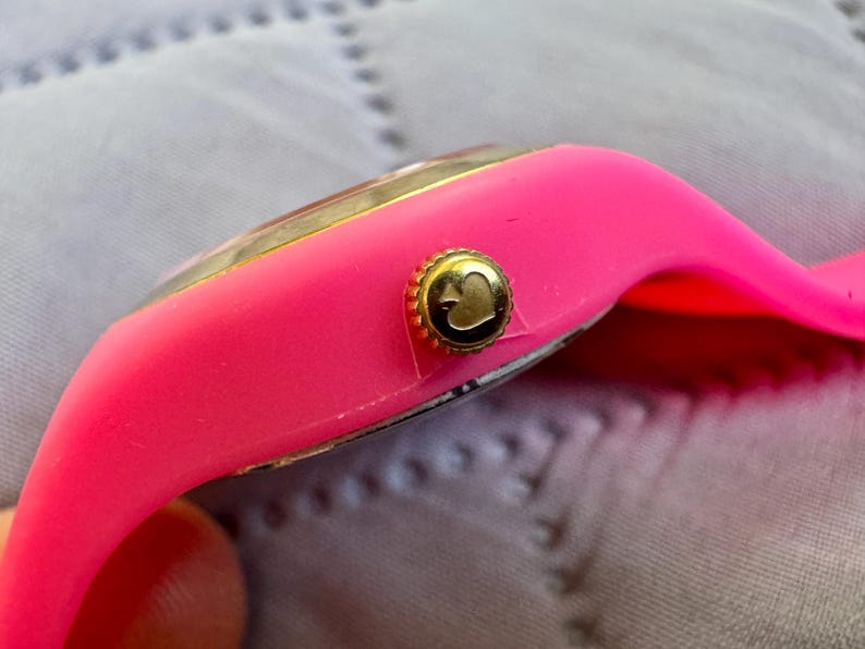 May include: Close-up of a bright pink wristwatch with a gold-colored bezel and crown. The crown features a heart-shaped design. The watch band is made of a flexible, pink material.