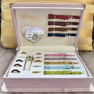 May include: A light pink rectangular watch gift set box featuring the text Ashley Princess. The interior includes a heart-shaped mirror, a silver-tone watch with a white strap, multiple colorful interchangeable watch straps, and several rings.