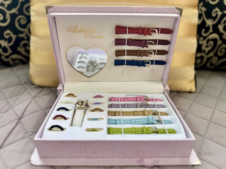 May include: A light pink rectangular watch gift set box featuring the text Ashley Princess. The interior includes a heart-shaped mirror, a silver-tone watch with a white strap, multiple colorful interchangeable watch straps, and several rings.