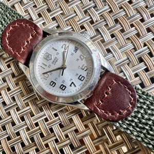 May include: A Swiss Army wristwatch with a silver-toned circular dial, black Arabic numerals, and a date window. The watch features a green woven fabric strap accented with brown leather tabs at the lugs, secured by visible stitching on the leather.