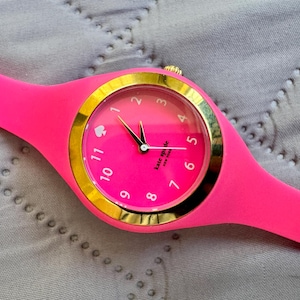 May include: A bright pink wristwatch with a gold-colored bezel and a matching pink band. The watch face is pink with gold numerals and hands. The brand name "Kate Spade" is printed on the watch face.