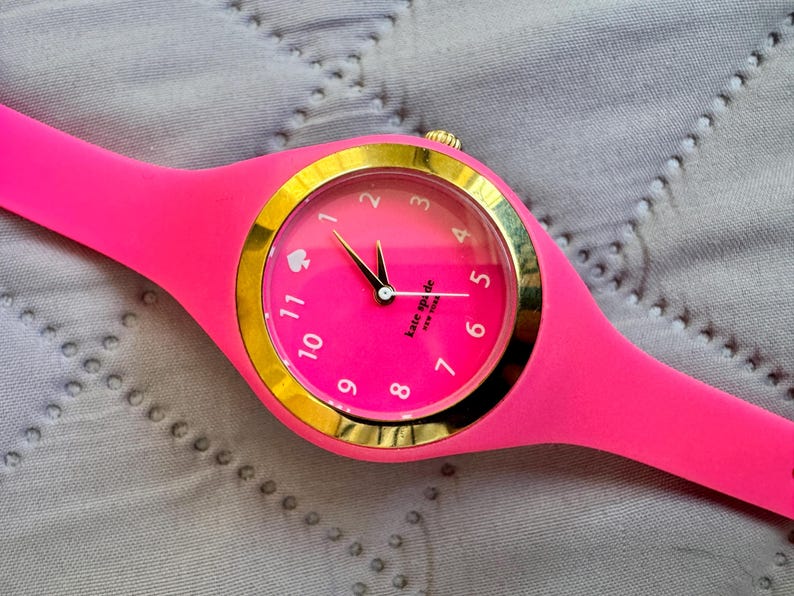 May include: A bright pink wristwatch with a gold-colored bezel and a matching pink band. The watch face is pink with gold numerals and hands. The brand name "Kate Spade" is printed on the watch face.