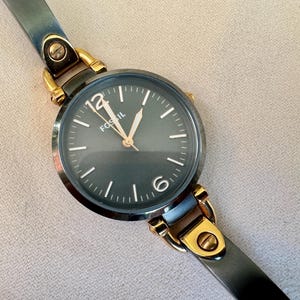 May include: A Fossil brand wristwatch with a dark gray face and gold-tone accents. The watch features a dark gray leather strap and gold-tone metal clasps. The watch face displays the brand name and the numbers 12 and 6.