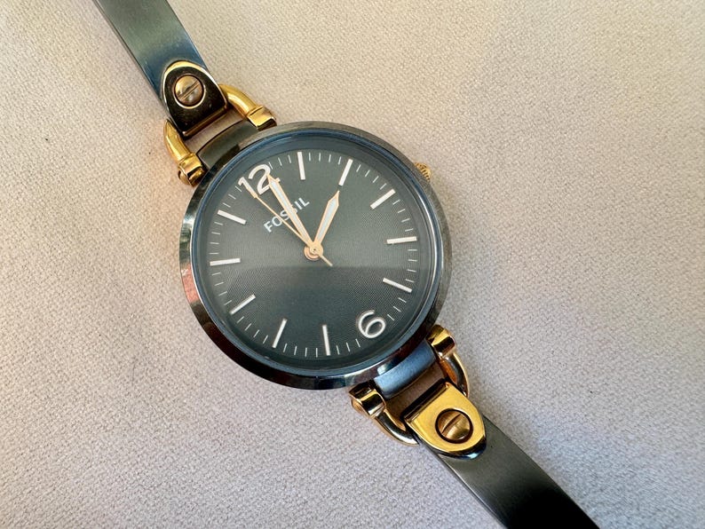 May include: A Fossil brand wristwatch with a dark gray face and gold-tone accents. The watch features a dark gray leather strap and gold-tone metal clasps. The watch face displays the brand name and the numbers 12 and 6.