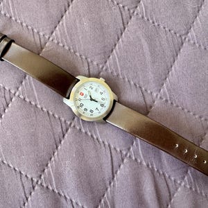 K&ouml;nnte beinhalten: Eine Armbanduhr mit rundem, silberfarbenem Geh&auml;use und wei&szlig;em Zifferblatt mit schwarzen arabischen Ziffern, schwarzen Zeigern und einem kleinen roten Logo auf der Zw&ouml;lf-Uhr-Position. Die Uhr hat ein glattes, dunkelbraunes Lederarmband mit Silberschlie&szlig;e.