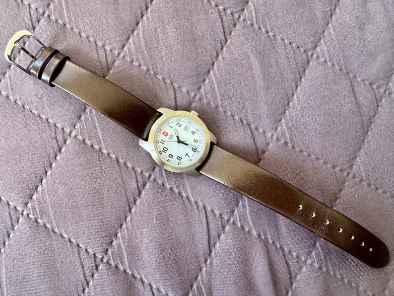 K&ouml;nnte beinhalten: Eine Armbanduhr mit rundem, silberfarbenem Geh&auml;use und wei&szlig;em Zifferblatt mit schwarzen arabischen Ziffern, schwarzen Zeigern und einem kleinen roten Logo auf der Zw&ouml;lf-Uhr-Position. Die Uhr hat ein glattes, dunkelbraunes Lederarmband mit Silberschlie&szlig;e.