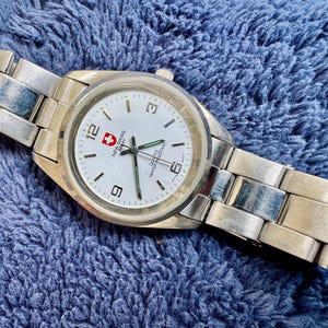 May include: A silver-toned wristwatch with a white face, featuring black numerals and hands, and the text "NEW SWISS ARMY". The watch has a metal band and is set against a blue textured background.