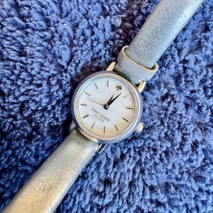 May include: A silver Kate Spade watch with a white face and a silver leather band. The watch face has black hands and a small spade logo. The band is approximately 0.5 inches wide.