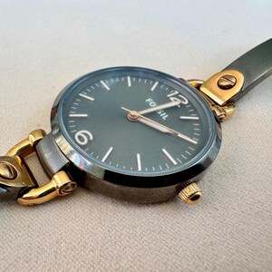 May include: A Fossil brand wristwatch with a dark gray face and gold-tone accents. The watch features a round face with white markers and hands, and a gray metal band. The watch is a stylish accessory.