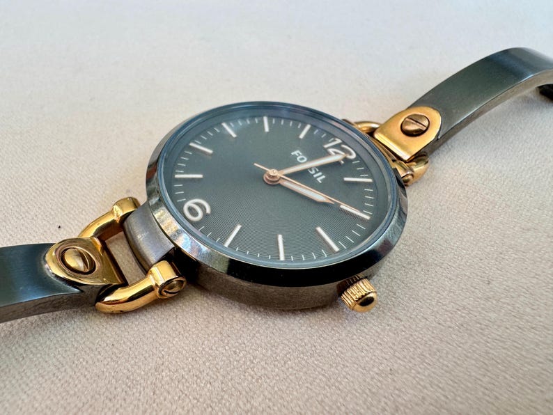 May include: A Fossil brand wristwatch with a dark gray face and gold-tone accents. The watch features a round face with white markers and hands, and a gray metal band. The watch is a stylish accessory.