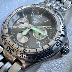 May include: A Fossil Blue analog wristwatch with a dark gray dial, silver-tone hands with luminous accents, and a stainless steel link bracelet. The watch face features multiple sub-dials, a date display, and a rotating bezel with engraved numbers.