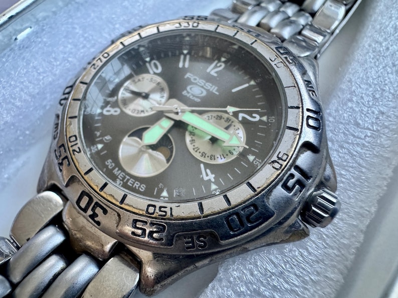 May include: A Fossil Blue analog wristwatch with a dark gray dial, silver-tone hands with luminous accents, and a stainless steel link bracelet. The watch face features multiple sub-dials, a date display, and a rotating bezel with engraved numbers.