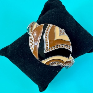 May include: A decorative bracelet with a geometric design in shades of brown, black, and cream, accented with small, sparkling crystals. The bracelet is set on a black velvet surface against a bright blue background.