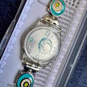 Może przedstawiać: Srebrny zegarek Swatch z przezroczystą plastikową obudową i wzorzystą tarczą. Pasek zegarka posiada turkusowe i białe okrągłe ogniwa. Tarcza zegarka wyświetla cyfry 12, 3 i 6. Zegarek jest w oryginalnym opakowaniu.