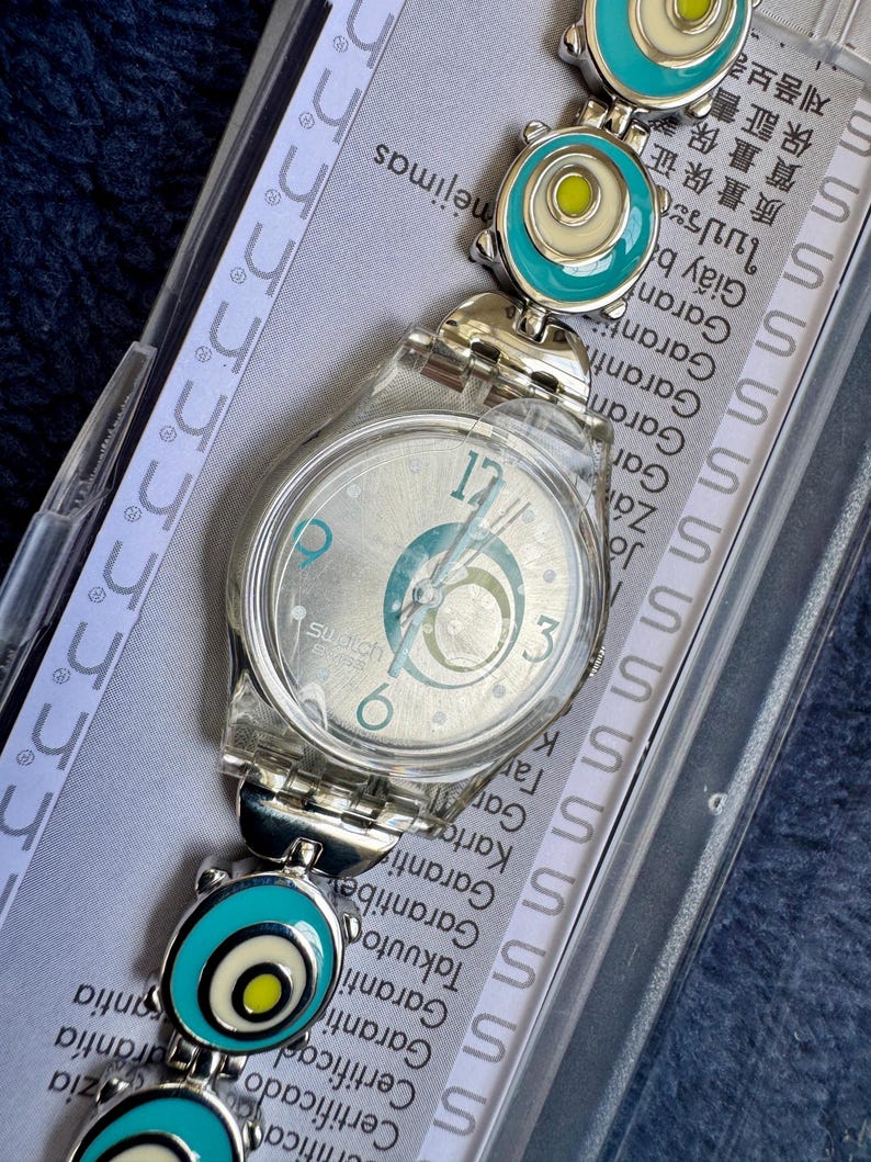 Może przedstawiać: Srebrny zegarek Swatch z przezroczystą plastikową obudową i wzorzystą tarczą. Pasek zegarka posiada turkusowe i białe okrągłe ogniwa. Tarcza zegarka wyświetla cyfry 12, 3 i 6. Zegarek jest w oryginalnym opakowaniu.