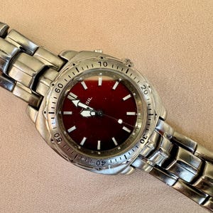 May include: A silver-toned Fossil watch with a burgundy face and a matching metal band. The watch features white hour markers and hands, with a date display. The watch is displayed on a neutral background.