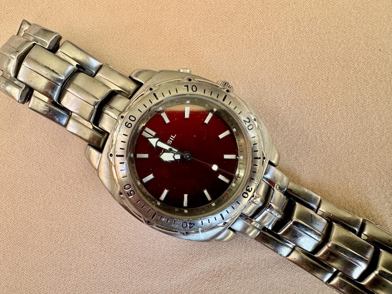 May include: A silver-toned Fossil watch with a burgundy face and a matching metal band. The watch features white hour markers and hands, with a date display. The watch is displayed on a neutral background.