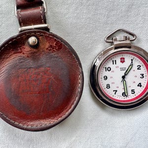 May include: A round brown leather case with a snap closure and a silver-tone Swiss Army pocket watch featuring a white dial, black numerals, a red inner ring, and luminous hands. The watch face displays the Swiss Army logo and text.