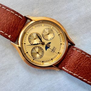 May include: A gold-tone Timex Quartz wristwatch featuring a round case, a textured brown leather strap with white stitching, and a gold dial. The face includes three sub-dials for day, date, and 24-hour time, along with a moon phase indicator.