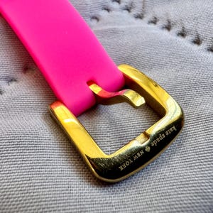 May include: Close-up of a bright pink watch strap with a gold-colored buckle. The buckle is engraved with "Kate Spade NEW YORK". The strap is made of a flexible material.
