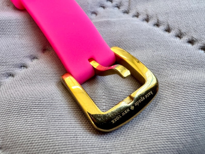 May include: Close-up of a bright pink watch strap with a gold-colored buckle. The buckle is engraved with "Kate Spade NEW YORK". The strap is made of a flexible material.
