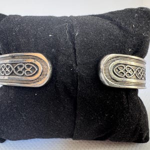 Puede incluir: Brazalete plateado con dise&ntilde;o abierto. El brazalete presenta un patr&oacute;n de nudo celta en relieve en la parte delantera. El brazalete se muestra sobre una superficie de terciopelo negro.